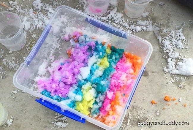 Clear plastic bin filled with snow dyed in vibrant rainbow colors including pink, orange, yellow, green, blue, and purple. Surrounding the bin are small cups used for coloring, with snow and splatters scattered on the concrete surface.