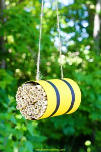 DIY bee hotel made from a painted tin can filled with cut bamboo tubes and hung from a tree with twine. The background is a blurred green garden setting.
