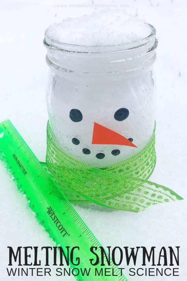 Clear mason jar filled with snow, decorated like a snowman with black dot stickers for eyes and mouth, an orange paper carrot nose, and a green glittery ribbon scarf wrapped around it. A green ruler lies beside the jar in the snow, used to measure the melting process. Text at the bottom reads “Melting Snowman Winter Snow Melt Science.”