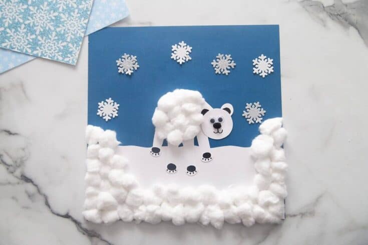 A winter-themed polar bear craft features a white bear made from paper and fluffy cotton balls for its body and snowy landscape. The background is decorated with silver snowflake stickers on blue paper, creating a cute and tactile Arctic scene for kids.