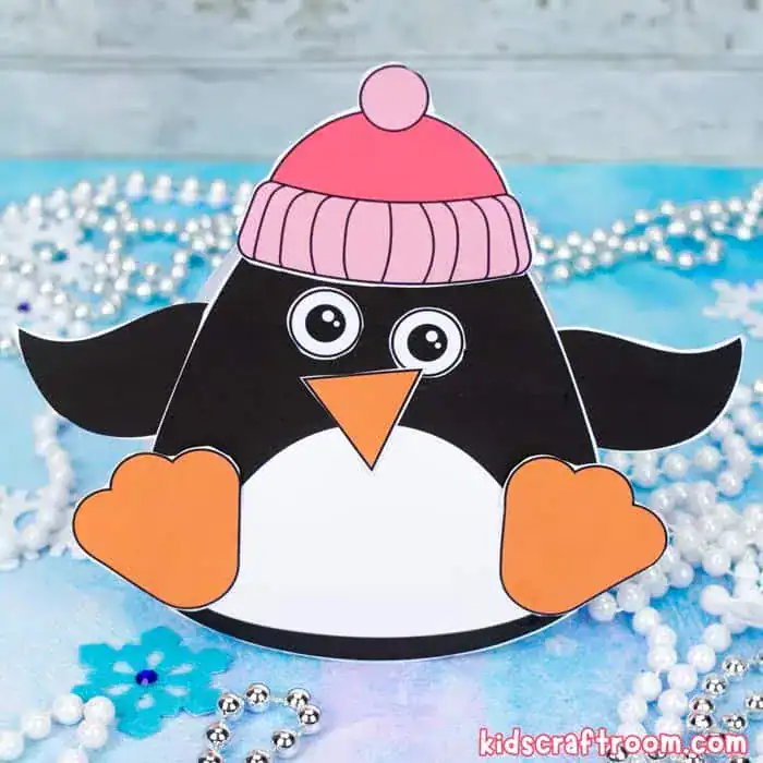 A cute paper penguin craft with large cartoon eyes, an orange beak and feet, and a pink winter hat sits on a blue surface decorated with snowflakes and silver beads. The cheerful design makes it ideal for a winter or holiday-themed kids' craft project.