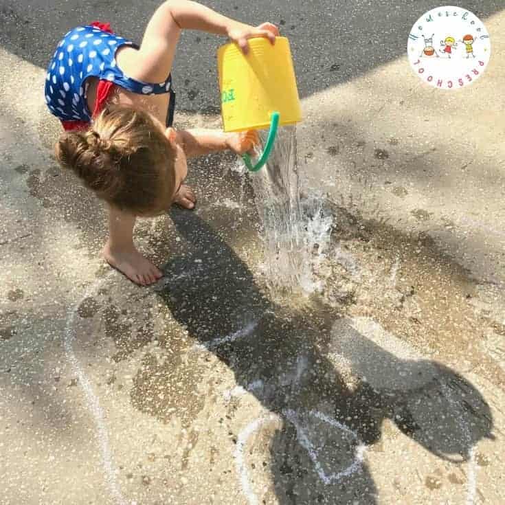 Child in summer pouring a bucket of water over some chalk writing in the pavement.