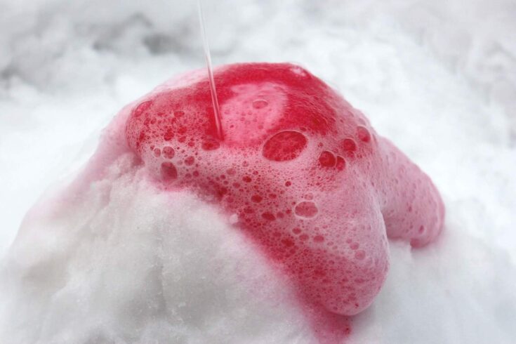 Red bubbly foam overflows from the top of a snow mound as liquid is poured onto it, demonstrating a snow volcano science experiment using baking soda and vinegar. The vibrant foam contrasts against the white snow background.