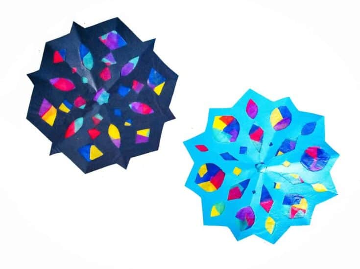 Two colorful paper snowflake crafts made from black and blue construction paper, with cut-out shapes backed by bright tissue paper in reds, yellows, blues, and purples. The stained-glass effect creates a vibrant, winter-themed decoration that glows when placed against light.