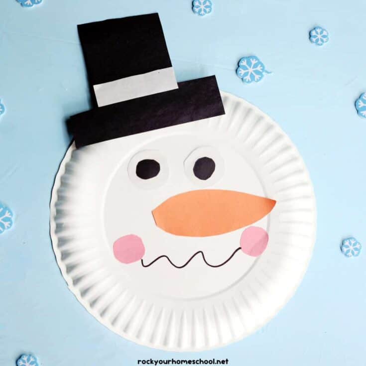 She Knows Alt Text said:A cheerful snowman face craft made from a white paper plate, featuring paper cutouts for a black top hat, round eyes, an orange carrot nose, pink cheeks, and a wavy black smile. The background is light blue with scattered paper snowflakes, creating a festive winter theme.