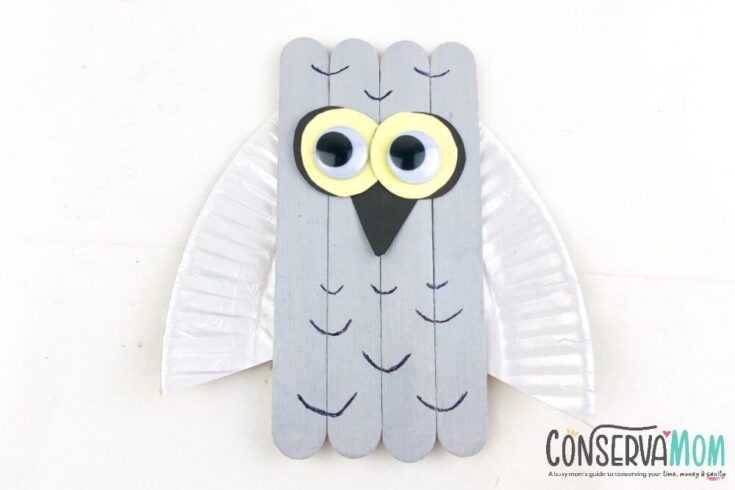 A cute paper penguin craft with large cartoon eyes, an orange beak and feet, and a pink winter hat sits on a blue surface decorated with snowflakes and silver beads. The cheerful design makes it ideal for a winter or holiday-themed kids' craft project.