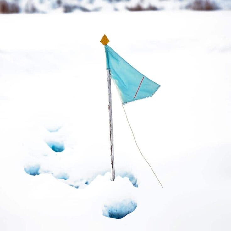 Small blue fabric flag with a red line and yellow tip attached to a wooden stick, standing upright in deep snow with visible footprints or holes around the base. The setup appears to be part of a winter wind or weather experiment.