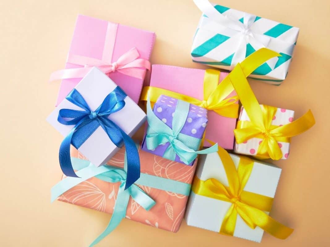 A colorful stack of gift boxes arranged on a light peach background, each wrapped in patterned or solid pastel paper with bright satin ribbons in blue, yellow, pink, and teal. The variety of sizes and vibrant colors gives a festive and cheerful vibe, perfect for birthdays, holidays, or celebrations.