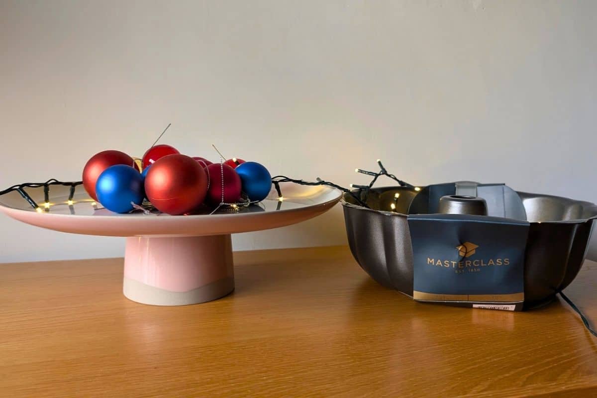 A pink pedestal cake stand decorated with red and blue Christmas baubles and string lights sits next to a MasterClass fluted bundt cake tin. Both items are arranged on a wooden surface with a festive holiday feel.
