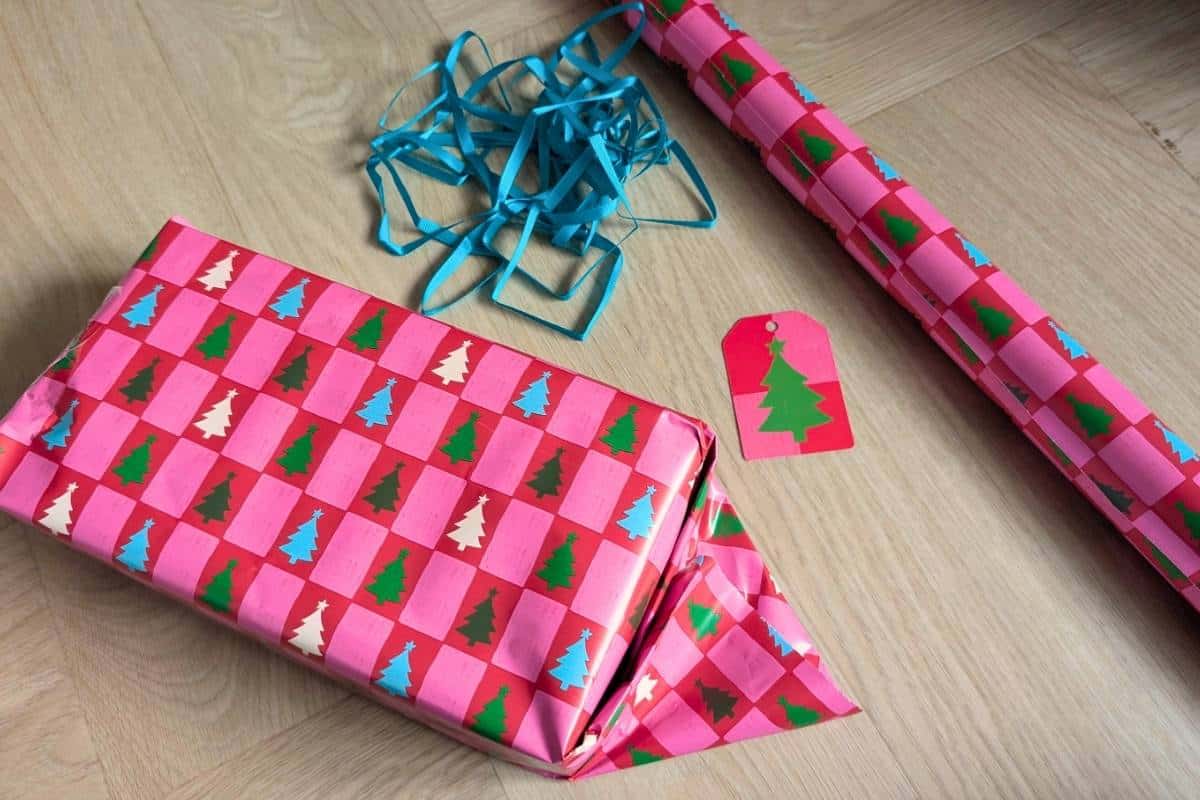 A partially wrapped Christmas gift sits on a wooden floor, covered in pink and red checkered wrapping paper with small Christmas trees in alternating colors. A matching roll of wrapping paper, a pile of blue ribbon, and a red gift tag with a green tree are nearby, ready for finishing touches.
