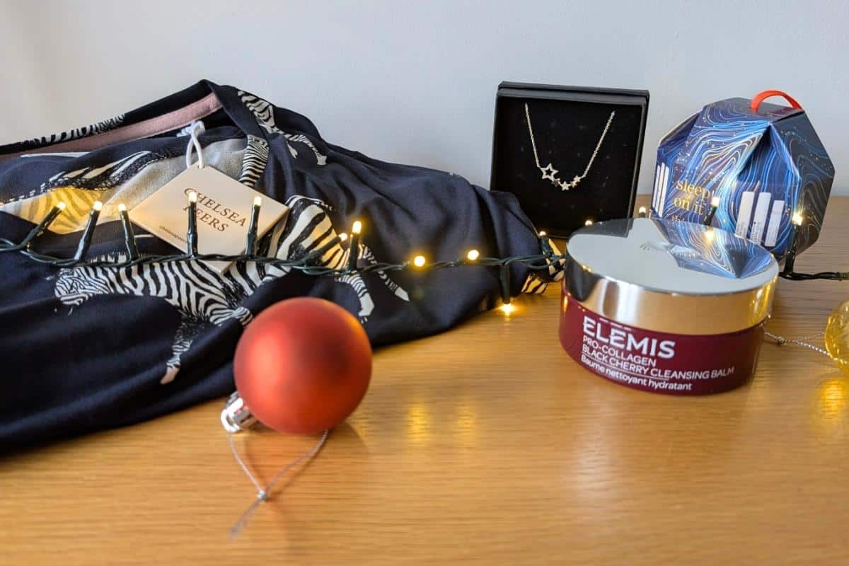 A flat lay of holiday gifts including a Chelsea Peers zebra-print loungewear set, a star-shaped pendant necklace in a black box, a jar of Elemis Pro-Collagen Black Cherry Cleansing Balm, and a “Sleep On It” nighttime skincare gift set. The scene is decorated with warm string lights and Christmas baubles on a wooden surface.