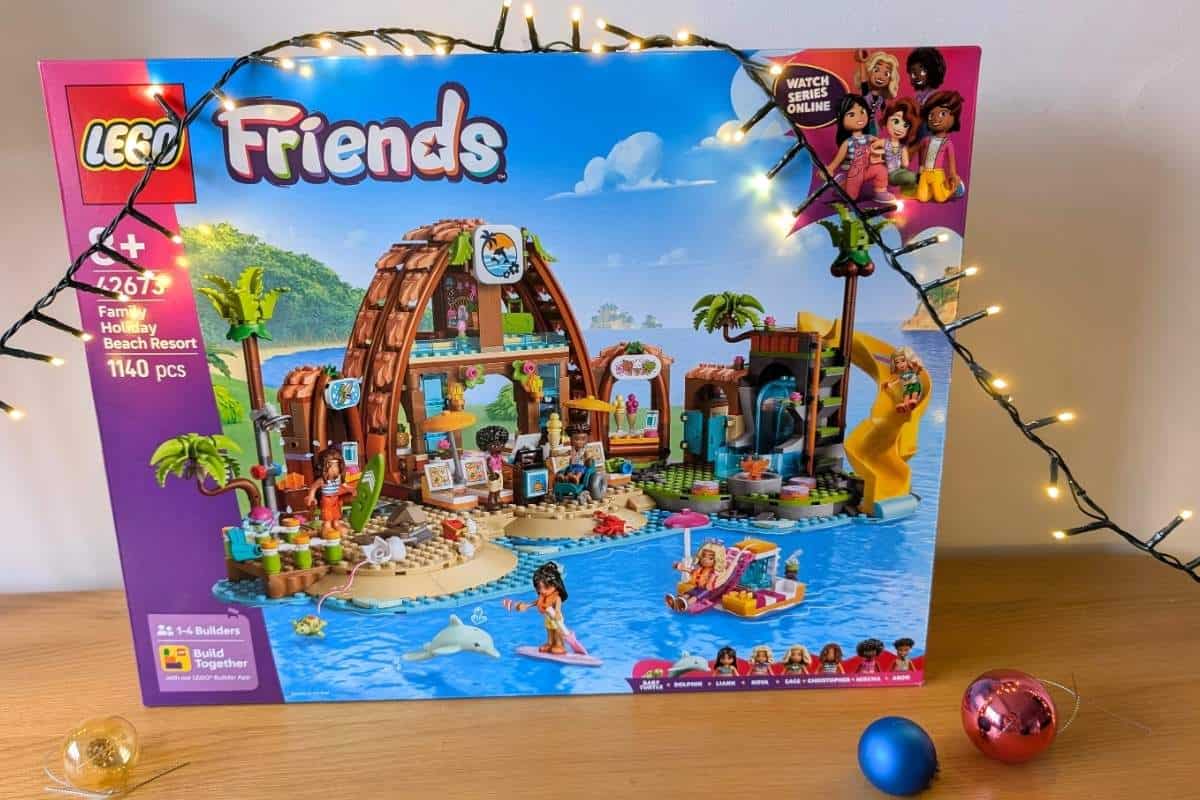 A LEGO Friends Family Holiday Beach Resort set displayed in front of a wall with glowing string lights and colorful Christmas ornaments. The box features a detailed beach resort scene with palm trees, water activities, and mini-figures enjoying the tropical setting.
