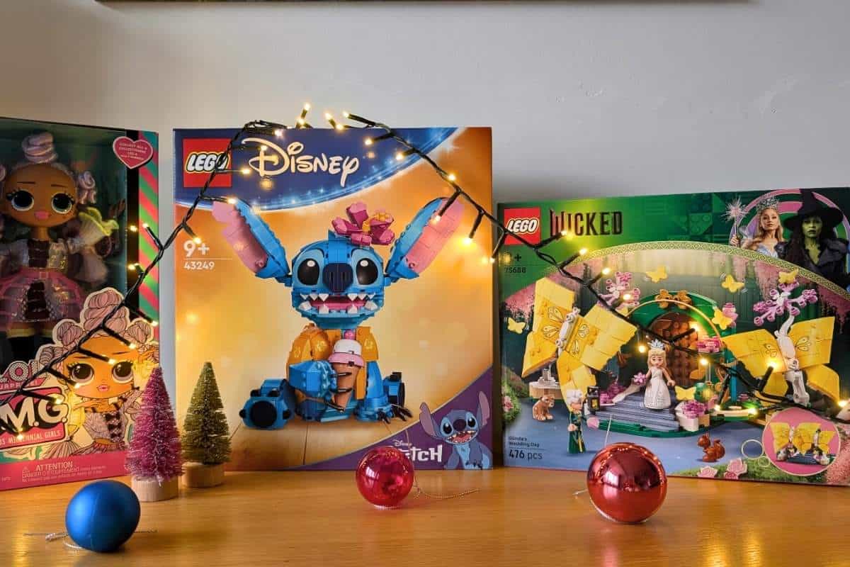 Three boxed toys displayed on a wooden surface, decorated with string lights and Christmas ornaments. The toys include an L.O.L. Surprise O.M.G. doll, a LEGO Disney Stitch set, and a LEGO Wicked set featuring Glinda and Elphaba.