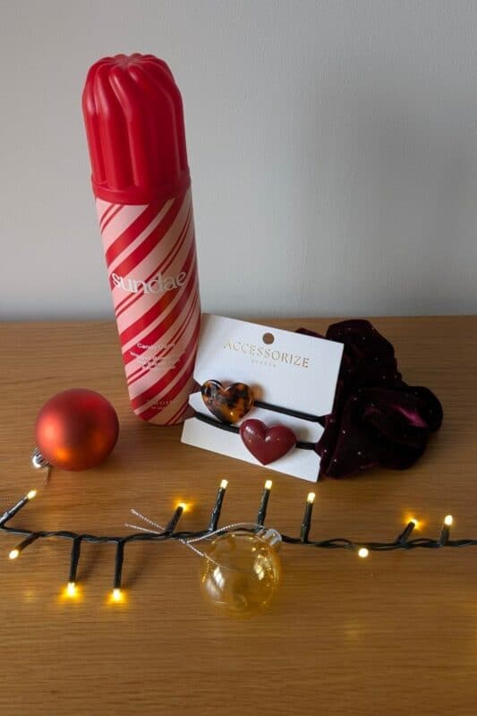 A festive gift set on a wooden surface featuring a red-and-white striped Sundae body wash shaped like a popsicle, a velvet scrunchie, and two heart-shaped hair ties from Accessorize. The scene is decorated with Christmas lights and baubles in red, gold, and clear tones.