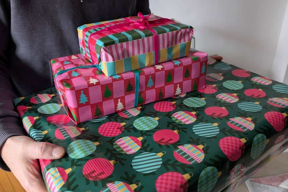 Three brightly wrapped Christmas presents stacked in someone’s arms, with each box featuring colorful holiday-themed wrapping paper. The top gift is wrapped in striped paper with a pink ribbon, the middle one has a red-and-pink checkered pattern with Christmas trees, and the bottom one is covered in green wrapping paper with multicolored ornament designs.