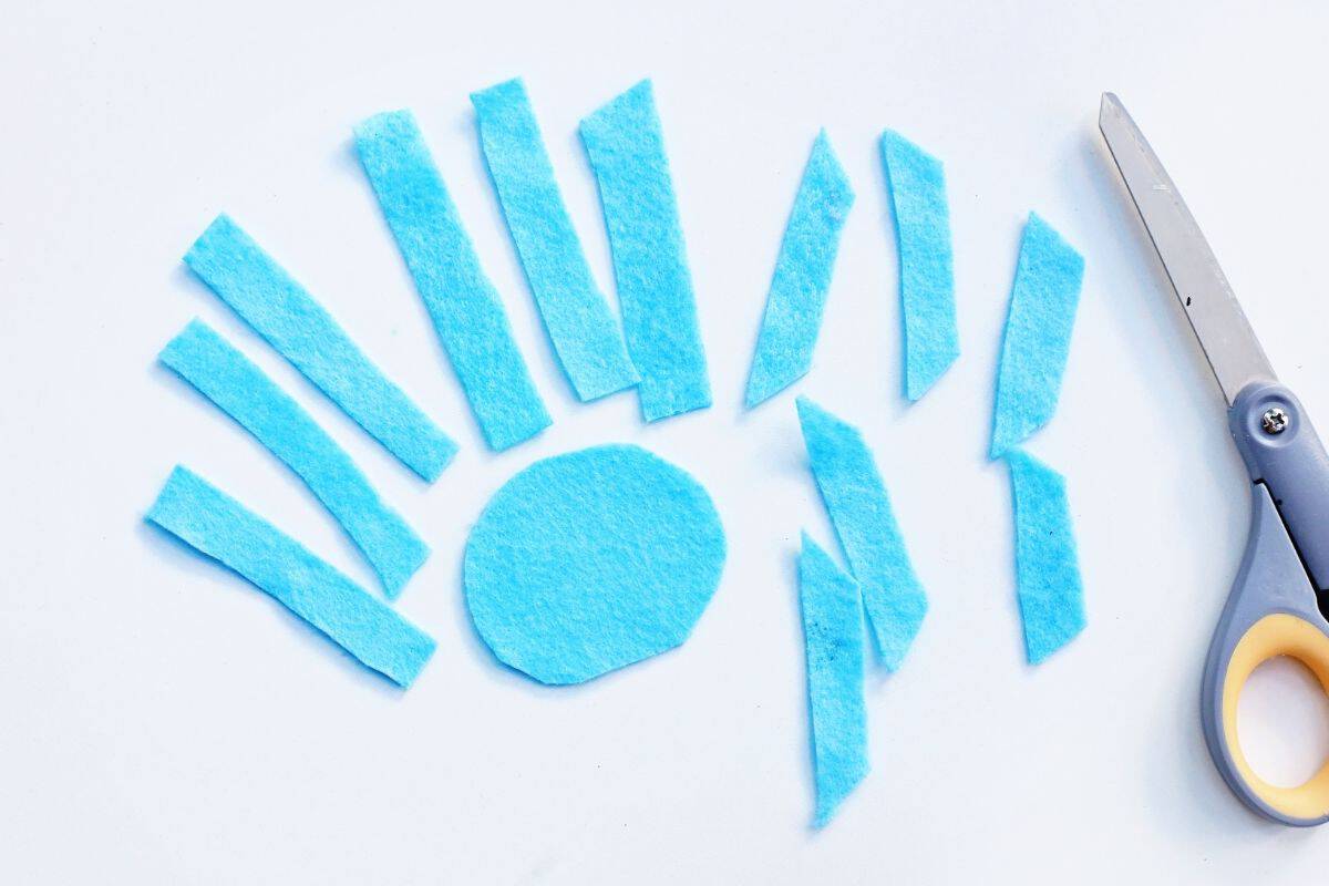 Various blue felt pieces are arranged on a white surface, including a central circle and multiple rectangular strips cut at different angles. A pair of scissors lies to the right, indicating a DIY craft in progress, likely a snowflake or similar shape.