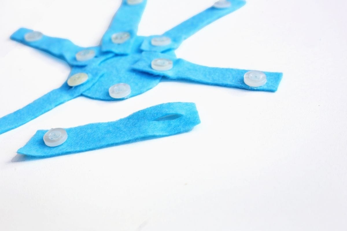 A close-up of a partially assembled felt snowflake craft shows blue felt strips with buttons attached to a central blue felt circle. One extra strip with a slit near its end lies separately, suggesting it's ready to be added to complete the design.