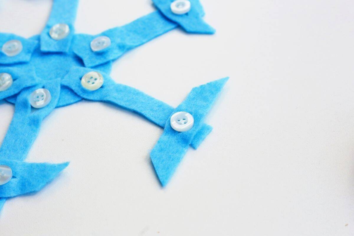 A close-up of a nearly finished felt snowflake craft shows blue felt arms radiating from a central circle, each decorated with a white button. One arm in focus has an added arrowhead-like felt piece, enhancing the snowflake's wintry design.