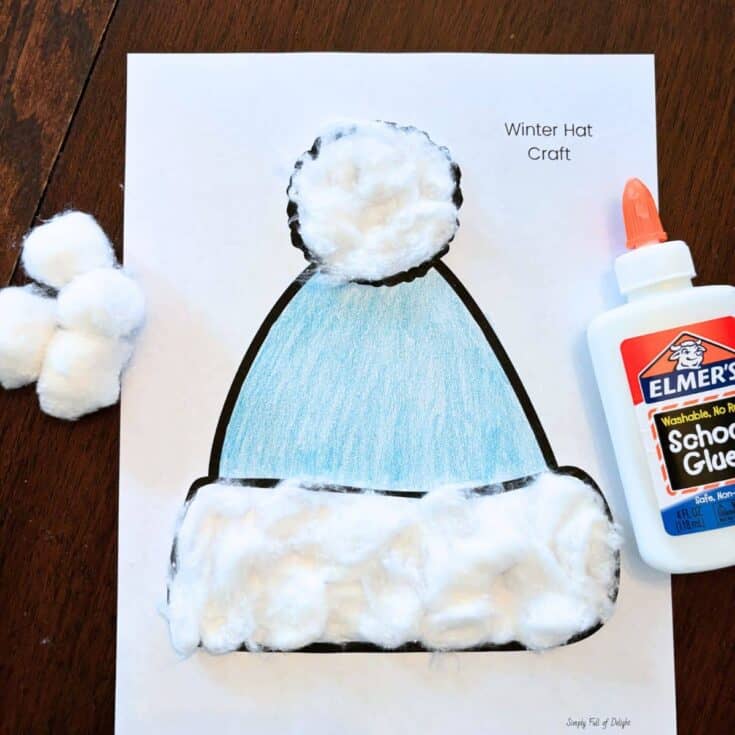 A simple winter hat craft featuring a blue crayon-colored hat outline on paper, decorated with glued-on cotton balls along the brim and pom-pom for a fluffy texture. A bottle of Elmer’s glue and extra cotton balls sit nearby, highlighting this as an easy, kid-friendly winter activity.