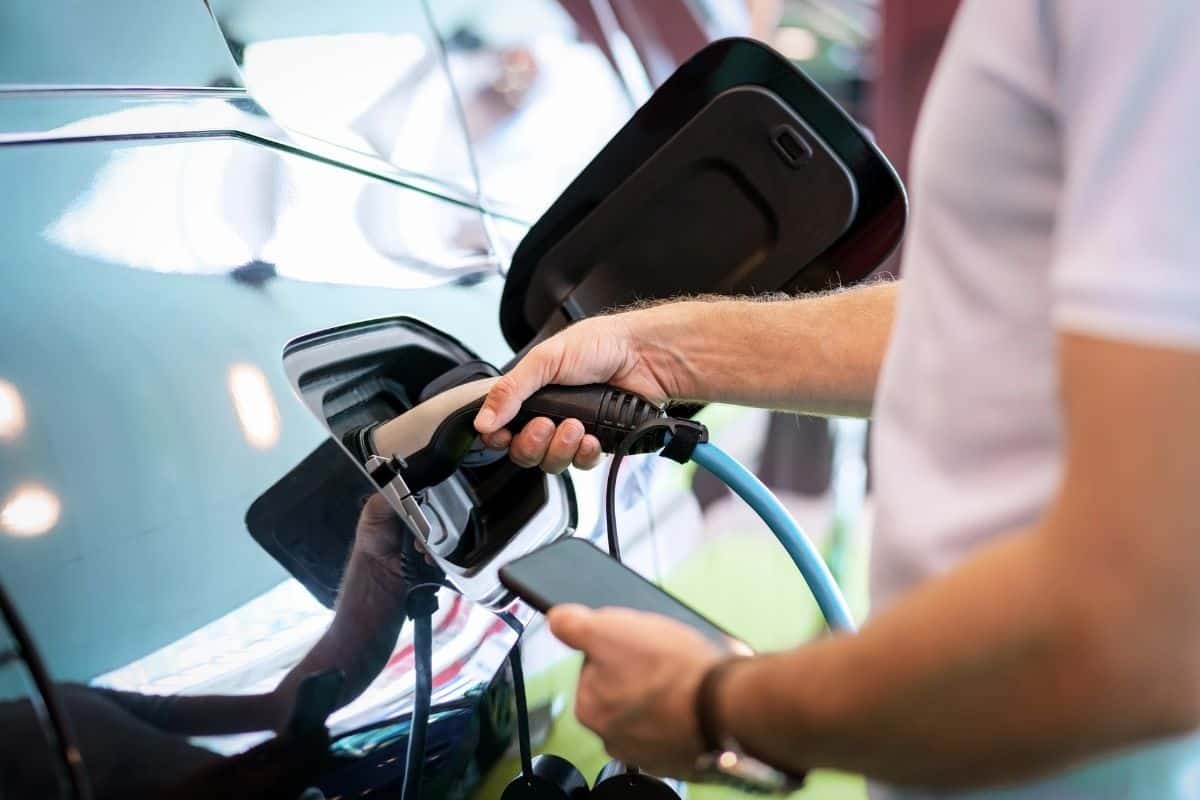 Close-up of a person plugging a charging cable into an electric vehicle. The individual holds a smartphone in one hand while connecting the charger with the other, with the car’s charging port open and visible.