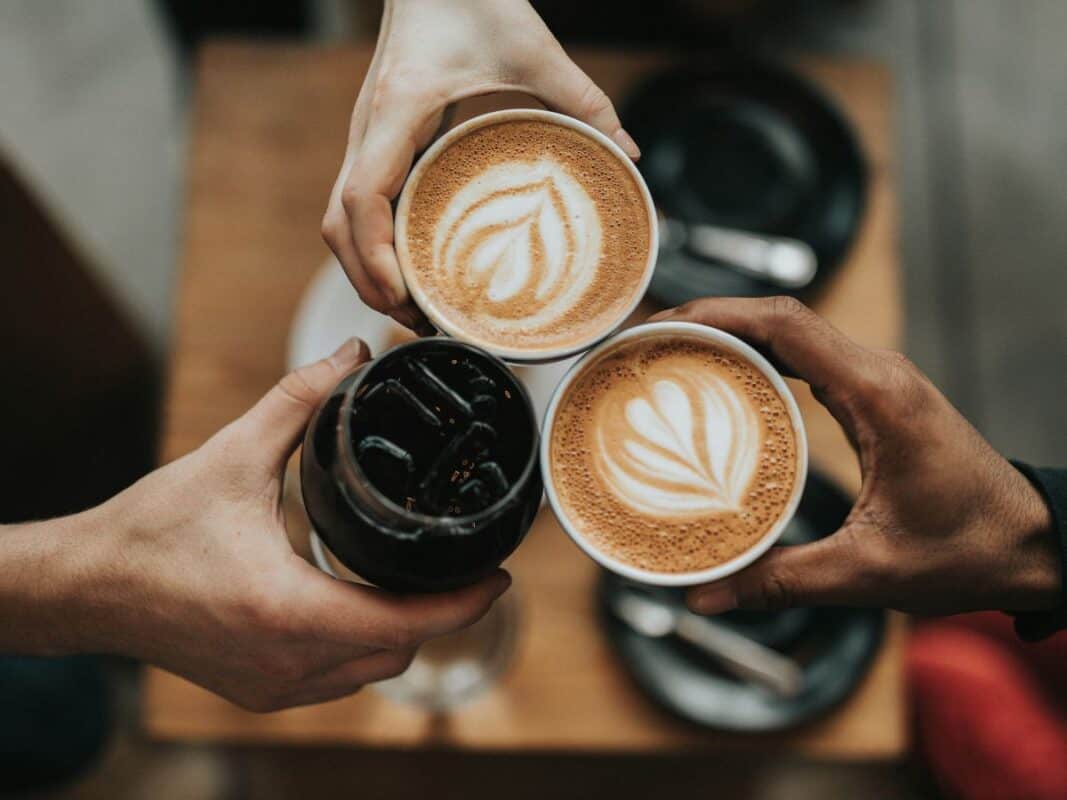 Three hands hold up drinks in a celebratory toast—two lattes with heart-shaped latte art and one glass of iced coffee. The drinks are set against a cozy café backdrop with blurred black saucers and a wooden table underneath.