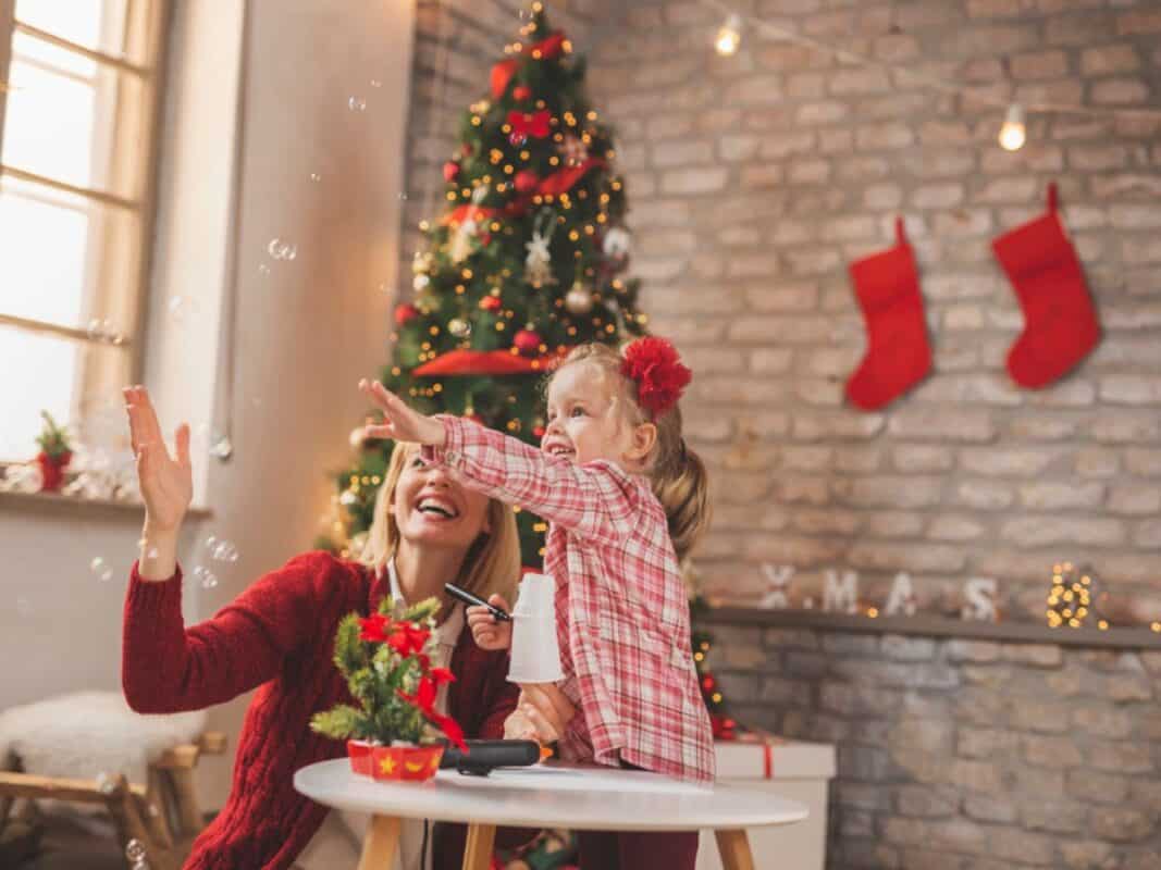 A mother and young daughter playfully reach for bubbles in a cozy, festively decorated room with a Christmas tree and red stockings hanging on a brick wall. The little girl holds a bubble wand while the mother smiles, both wearing red tones that match the holiday decor.