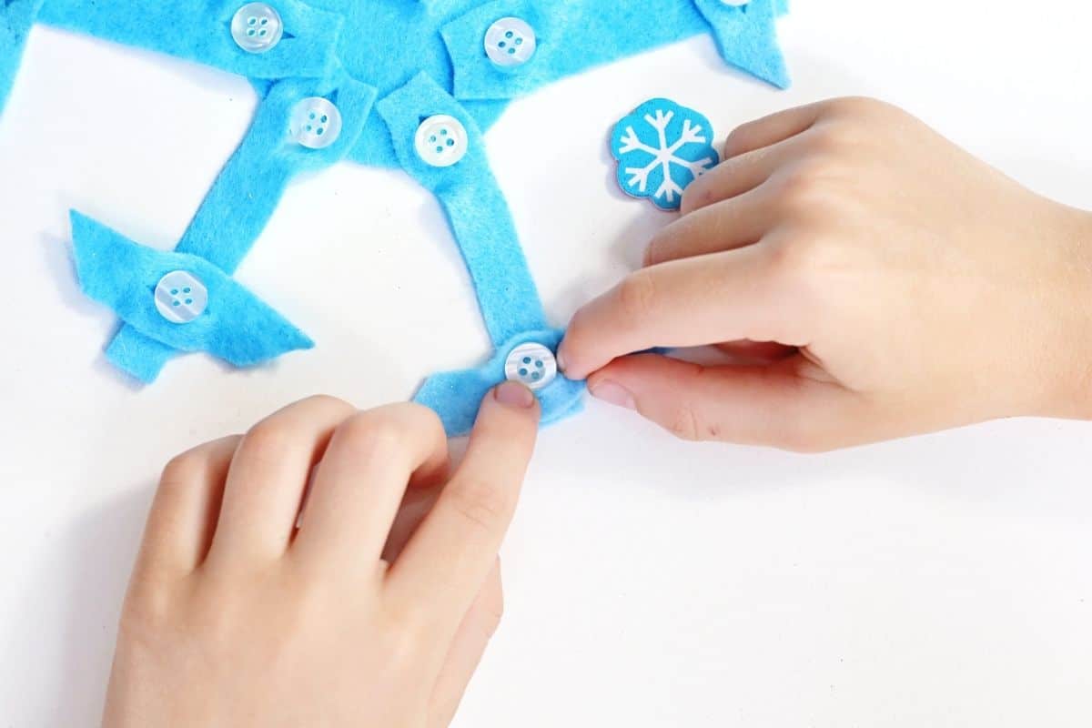 Two small hands are assembling a blue felt snowflake craft using white buttons as fasteners. A blue felt piece with a printed white snowflake design sits nearby on a white surface.
