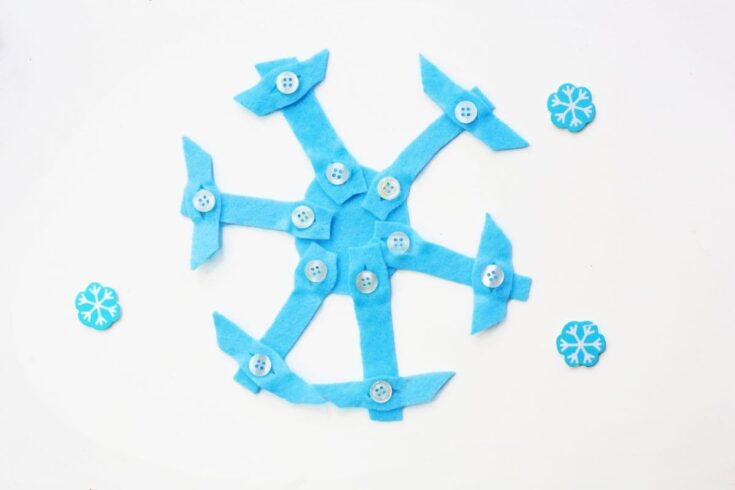 A handmade snowflake craft made from blue felt and white buttons arranged in a radial pattern on a white background. Three small blue snowflake embellishments are placed around it for decoration, giving it a winter-themed look.