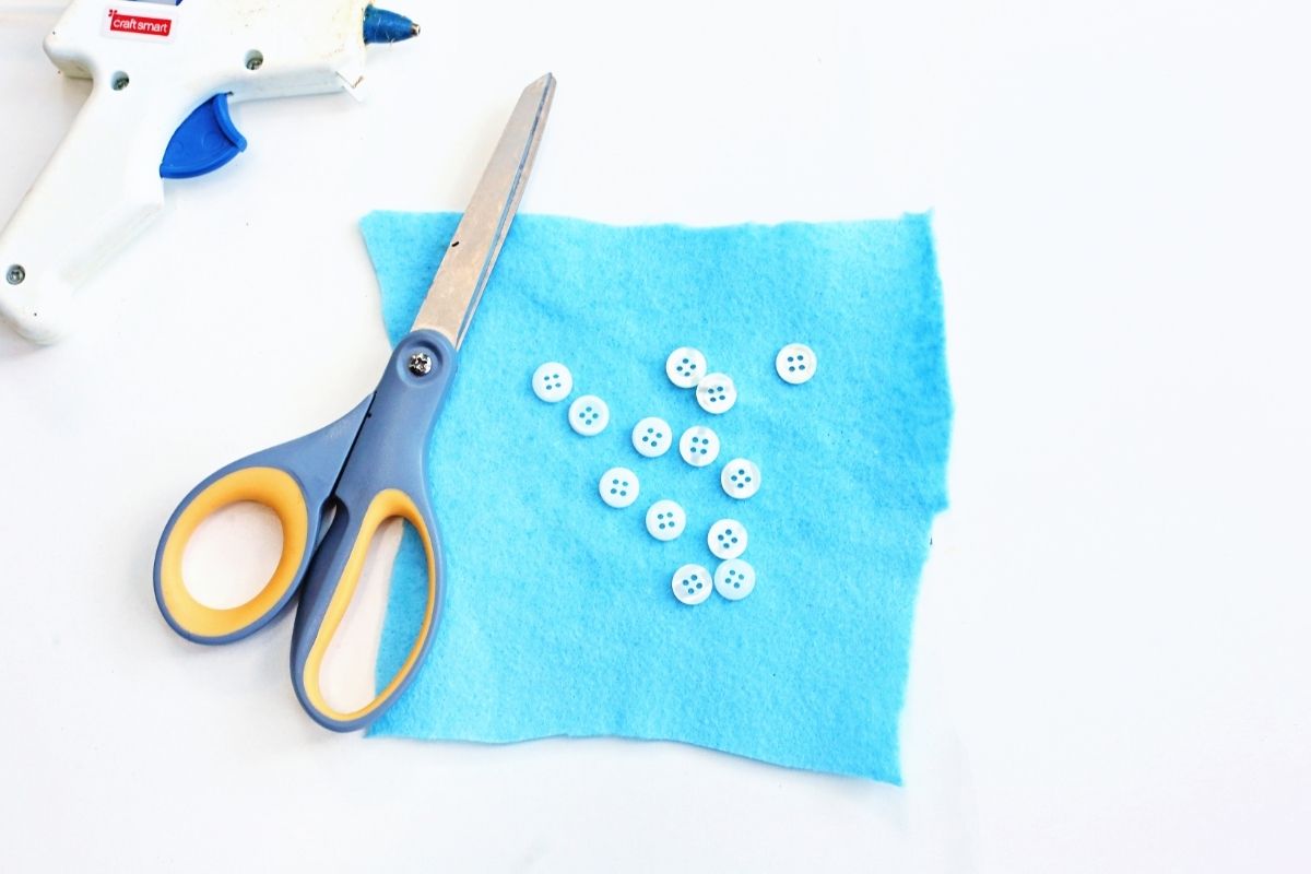 A pair of craft scissors, a white hot glue gun, and a square piece of blue felt are laid out on a white surface. Small white buttons are scattered on the felt, suggesting a sewing or DIY craft project in progress.