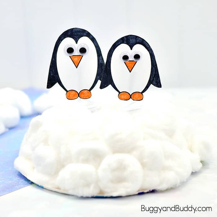 Two paper penguin crafts with googly eyes, orange beaks, and orange feet are perched on a mound of white cotton balls, simulating snow. The background and setting create a wintry scene, perfect for a kid-friendly Arctic or winter animal project.