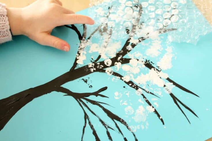A child’s hands press tissue or plastic wrap onto a painting of a tree with black branches on blue paper, creating a textured effect that mimics white blossoms or snow. The artwork captures a seasonal craft activity, likely representing spring or winter.