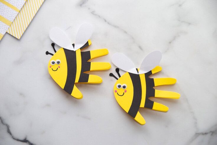 Two handprint bee crafts made from yellow, black, and white paper are shown on a white surface, with decorative yellow-striped and polka dot paper in the background. Each bee has a smiling face with googly eyes, pink cheeks, white heart-shaped wings, and black stripes, designed to resemble a child’s handprint.