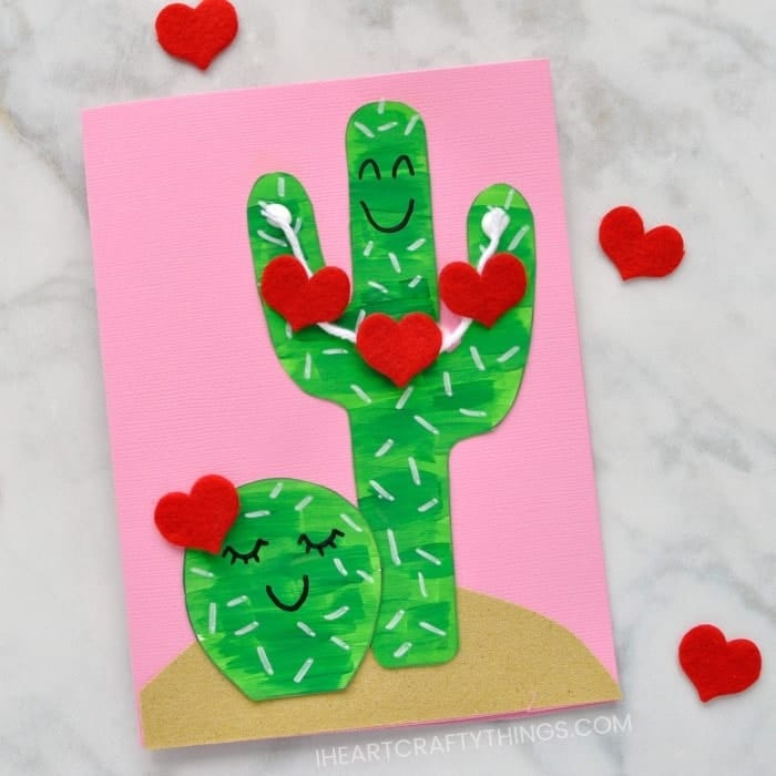 A handmade Valentine’s card features two green paper cacti with white painted spines and smiling faces, set against a pink background with a tan paper ground. Red felt hearts are attached to the cacti and scattered around, adding a playful and affectionate touch to the desert-themed craft.