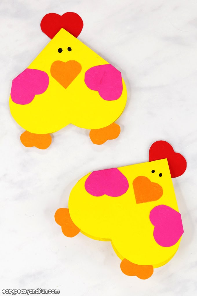 Two heart-shaped chicken crafts made from colorful paper are displayed on a white surface, featuring bright yellow bodies, pink wings, orange beaks and feet, and red heart combs. Each chicken has black dot eyes and is designed using multiple heart shapes, making them a festive and fun Valentine’s Day activity for kids.