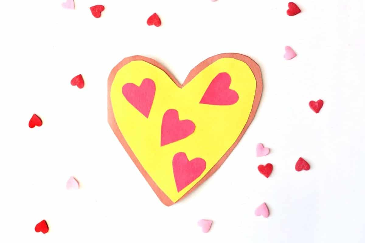 A handmade heart-shaped Valentine card made to look like a pizza, with a brown crust, yellow center, and red heart-shaped “pepperoni” pieces. The card is surrounded by scattered red and pink heart confetti on a white background.