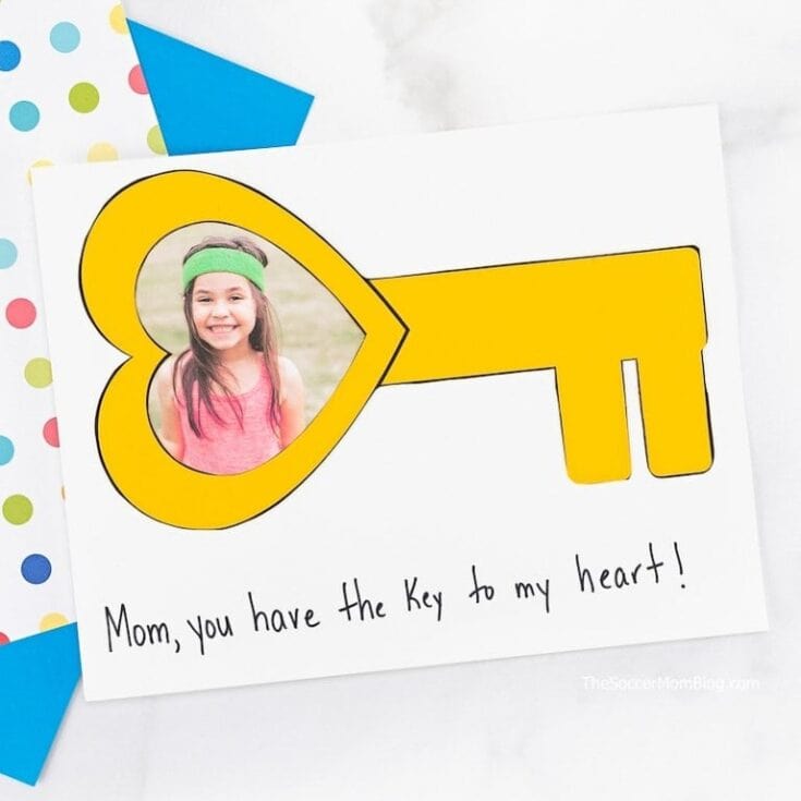 A handmade card features a large yellow key with a heart-shaped bow framing a photo of a smiling child, along with the handwritten message, "Mom, you have the key to my heart!" beneath it. The card is set against a white background with polka dot and blue paper accents, making it a sweet and personal DIY Mother’s Day or Valentine’s Day craft.