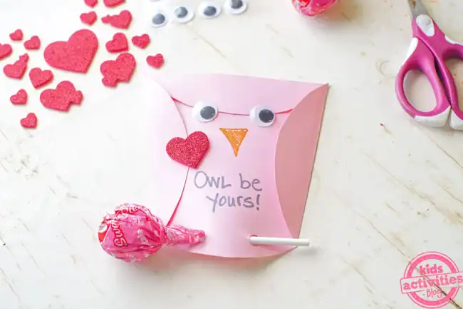 A pink owl-shaped Valentine made from folded paper features googly eyes, a heart-shaped nose, and the handwritten message “Owl be yours!” on its belly. A lollipop is tucked into the bottom as a treat, surrounded by heart stickers, craft supplies, and scissors on a white tabletop.
