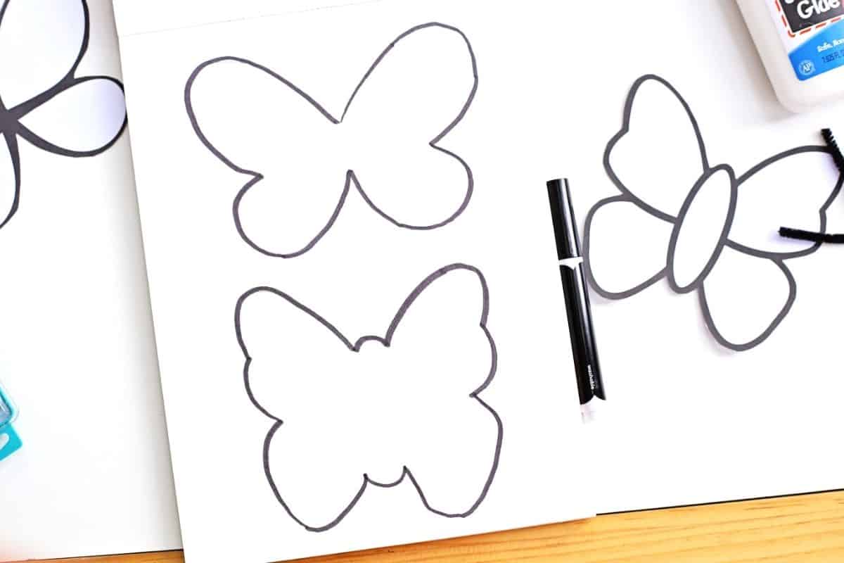 Two black-outlined butterfly templates are drawn on a white sheet of paper, with one completed butterfly cutout to the right and crafting supplies like glue and a black marker nearby.