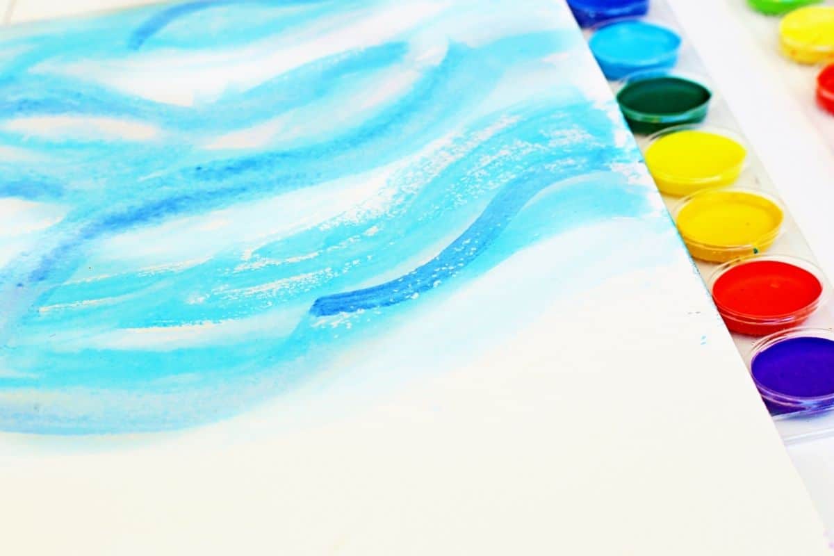 Close-up of a watercolor painting with swirling shades of blue on white paper, mimicking a winter sky or snowdrift pattern. A watercolor palette with bright colors, including red, yellow, green, and purple, is partially visible along the edge.