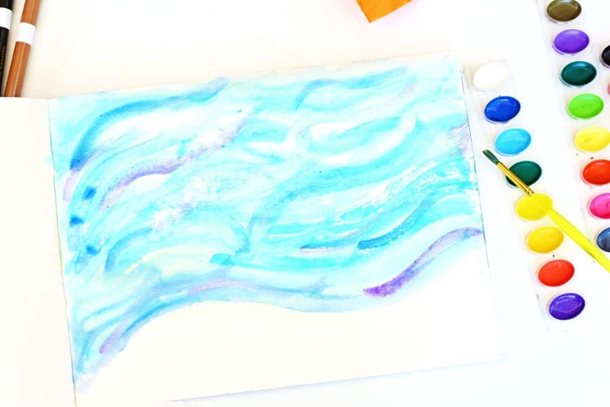 Watercolor painting featuring flowing blue and purple strokes on white paper, creating a dreamy winter sky effect. A watercolor paint set with a yellow-handled brush lies beside the paper, and two markers are partially visible in the top left corner.