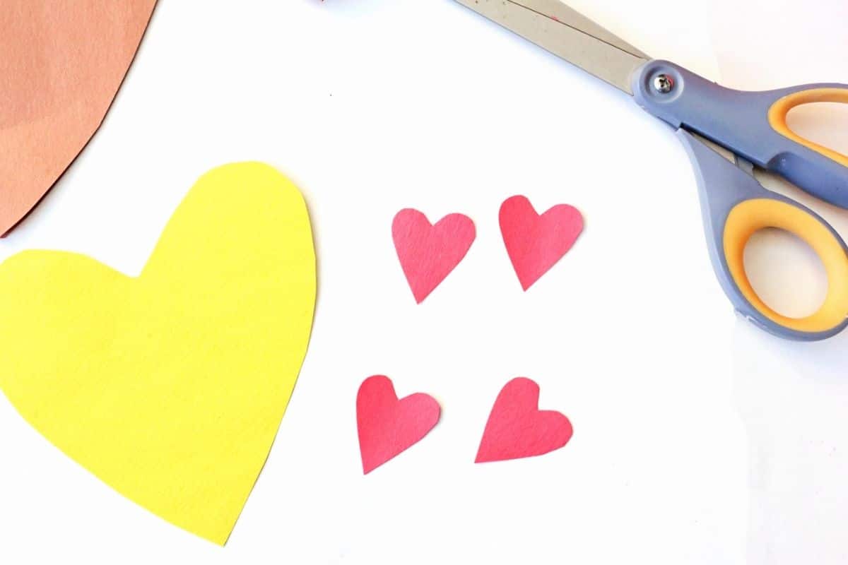 A yellow heart and four small red heart cutouts made from construction paper are arranged on a white surface, next to a pair of scissors and part of a brown heart shape, showing pieces for assembling a Valentine heart pizza card.