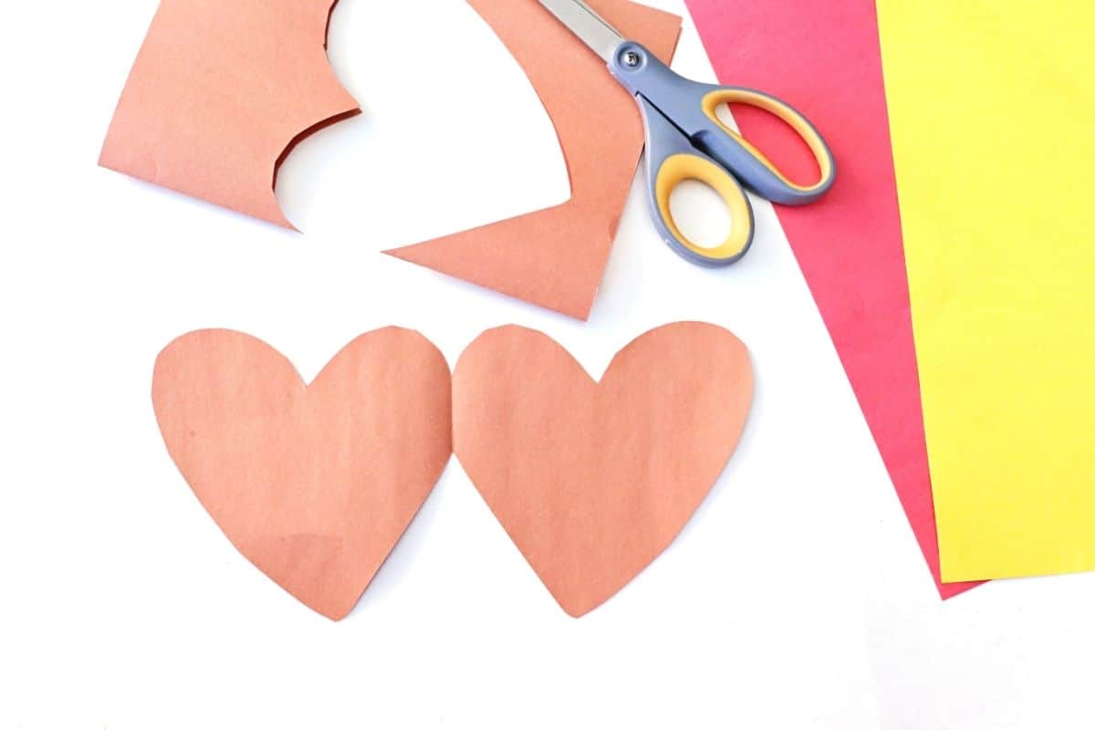 Two brown paper hearts cut out from construction paper lie on a white surface, next to a pair of scissors and sheets of red and yellow paper. One heart has just been cut from a larger sheet, showing the crafting process for a handmade Valentine card.