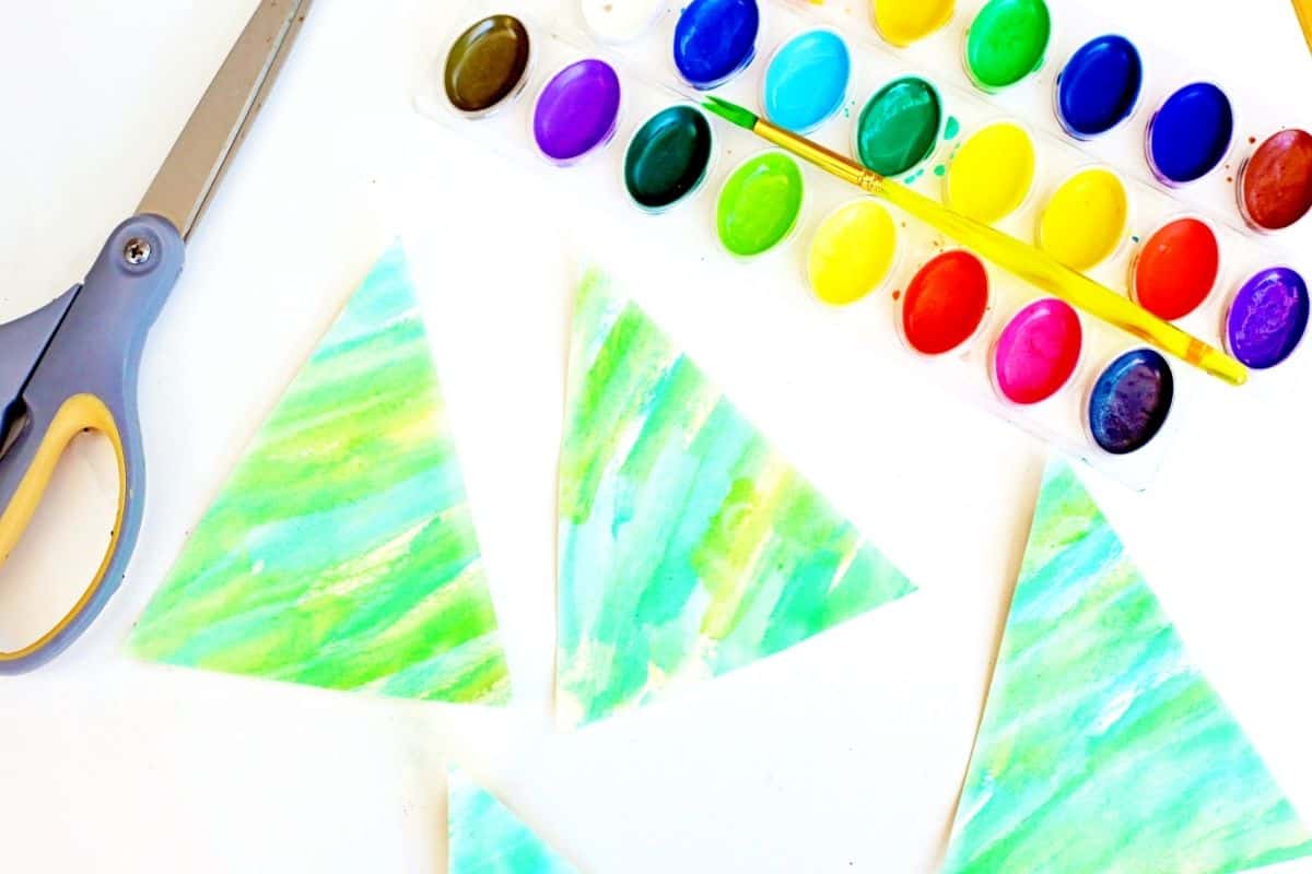 Three green watercolor-painted triangles cut from paper, arranged on a white surface to resemble tree shapes. A pair of scissors, a watercolor palette, and a yellow-handled brush are also visible, showing supplies used for a kids’ winter art project.