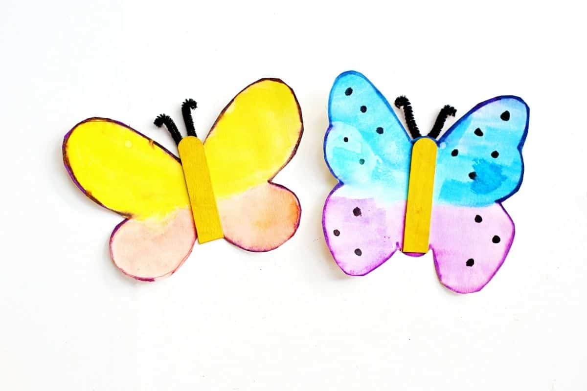 Two finished butterfly crafts made from watercolor-painted paper, each with a yellow-painted craft stick for the body and black pipe cleaner antennae. The left butterfly is yellow and orange, while the right one is blue and purple with black spots.