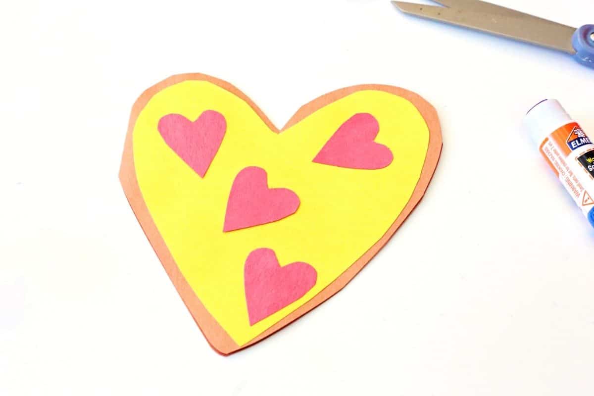 Completed heart pizza Valentine card made from construction paper, featuring a brown crust, yellow center, and four red heart-shaped toppings. A pair of scissors and glue stick are visible nearby, showing the finished result of the craft project.