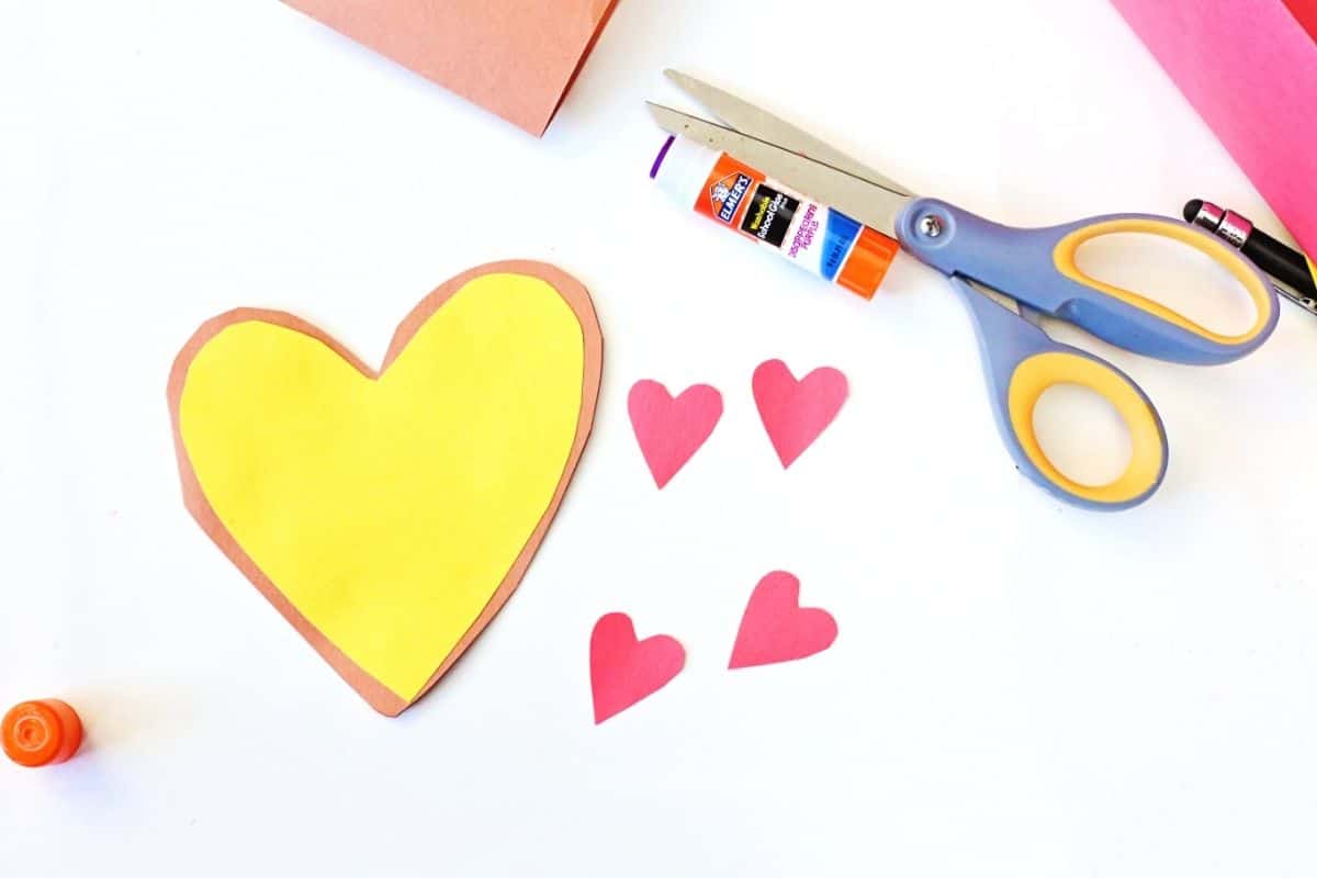 A partially assembled heart-shaped Valentine card made to look like a pizza sits on a white surface, with a yellow heart glued onto a brown heart base. Nearby are four red heart cutouts, scissors, a glue stick, and a pencil, showing the final crafting steps.
