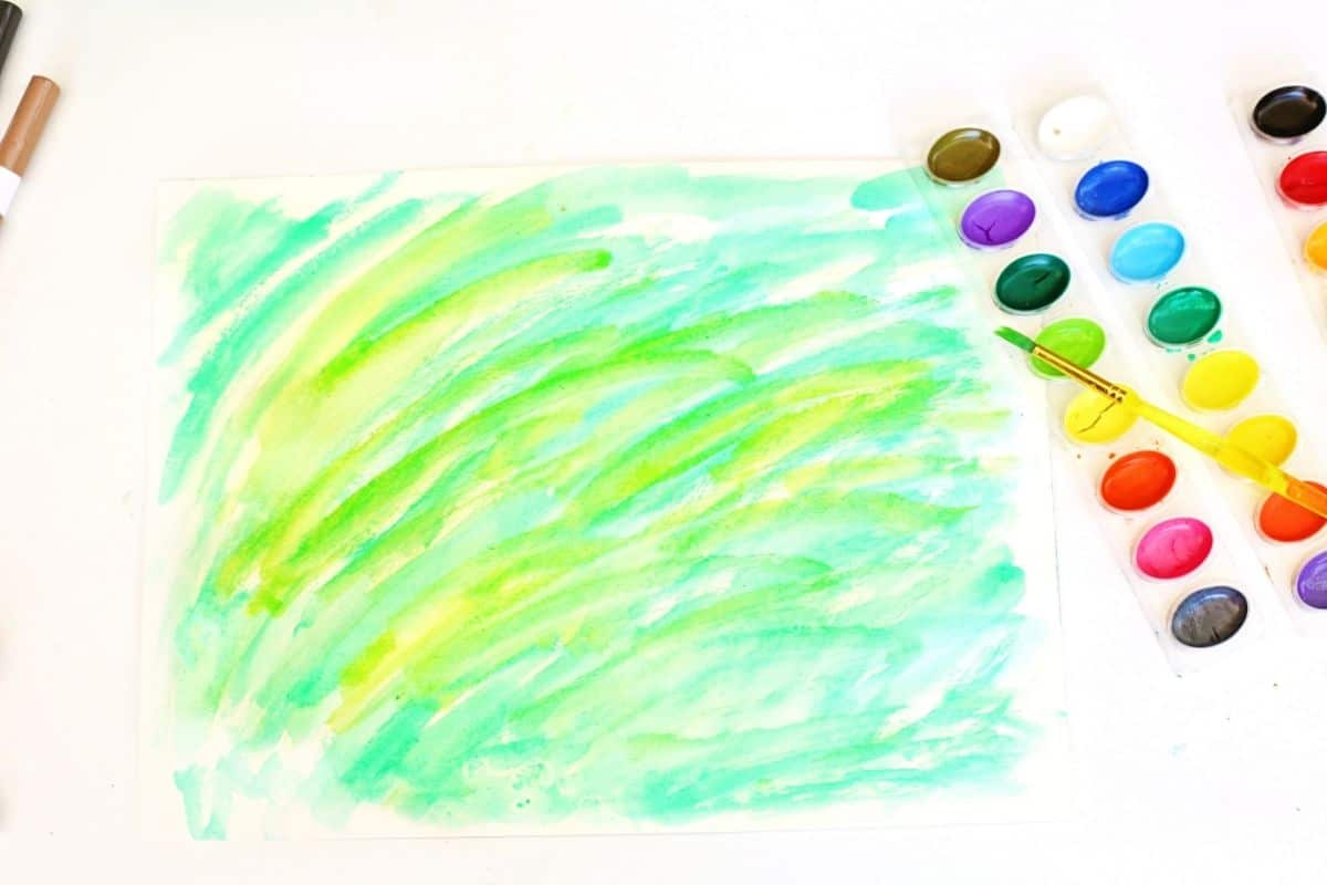 or forest background. A watercolor palette with a yellow-handled brush rests nearby, and two markers are partially visible on the left side.