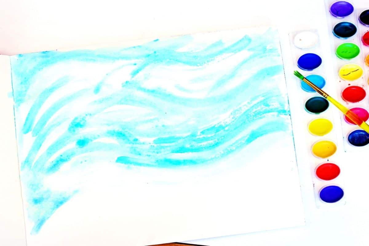 Watercolor painting in progress showing light blue wavy brush strokes across a white sheet of paper, resembling a wintry sky or snowy landscape. A yellow-handled paintbrush rests on a watercolor palette filled with vibrant paint colors on the right side.