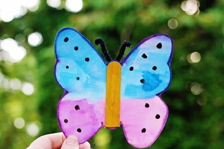 A hand holds up a finished butterfly craft against a blurred green outdoor background. The butterfly is painted in blue and purple with black dots, has a yellow craft stick body, and black pipe cleaner antennae.