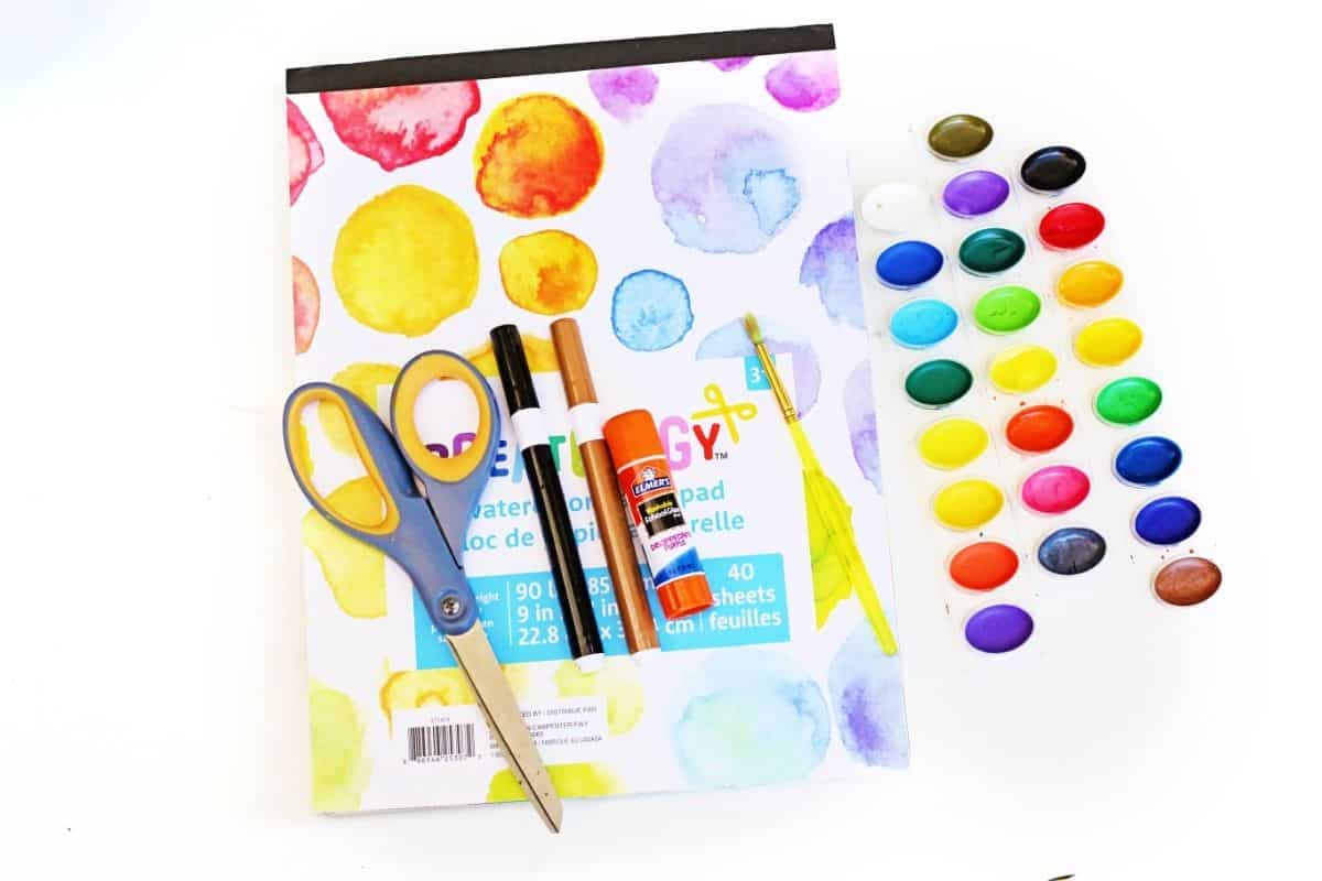 Flat lay of kids’ art supplies including a watercolor pad, a pair of scissors, black and brown markers, a glue stick, a yellow paintbrush, and a tray of colorful watercolor paints. The materials are arranged neatly on a white background, ready for a creative project.